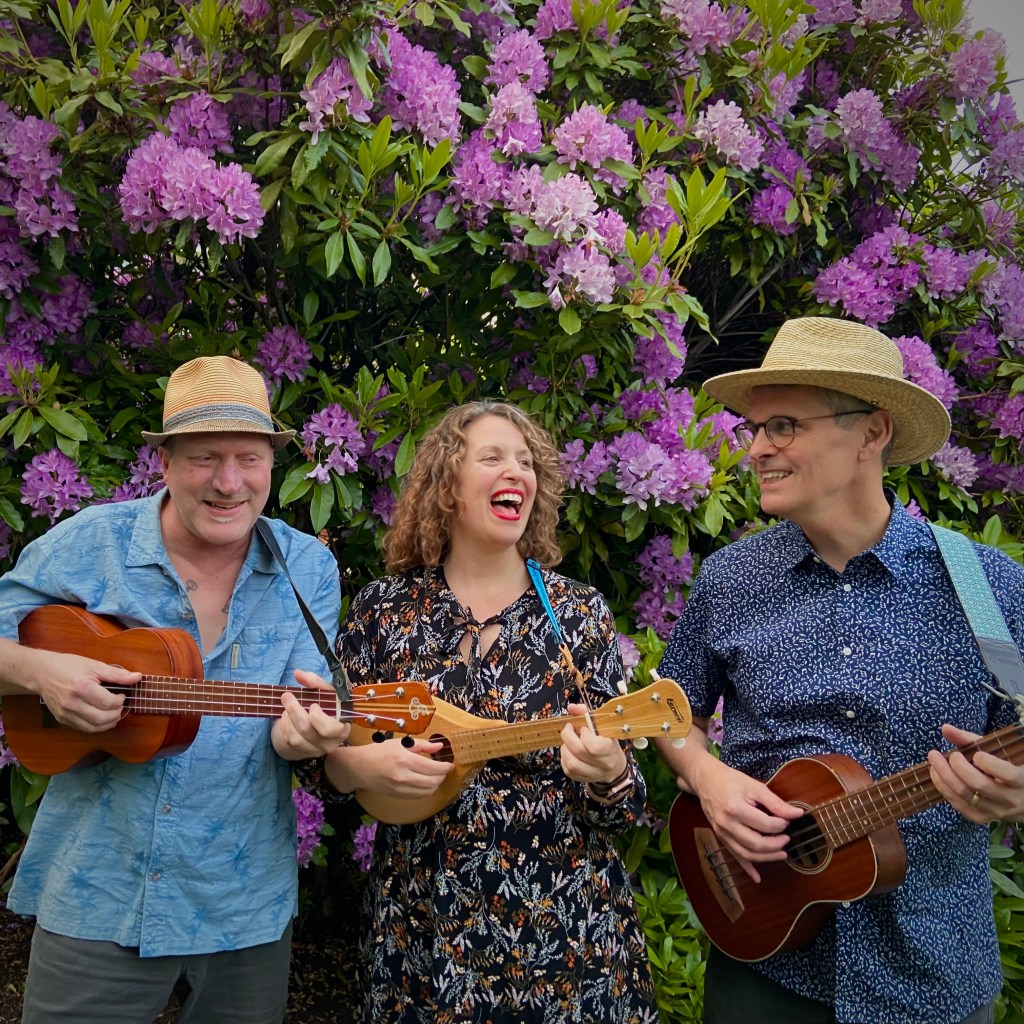 Michael, Sunga and Eric of the Honeyville Rascals laughing with their instruments in front of a large rhododendron